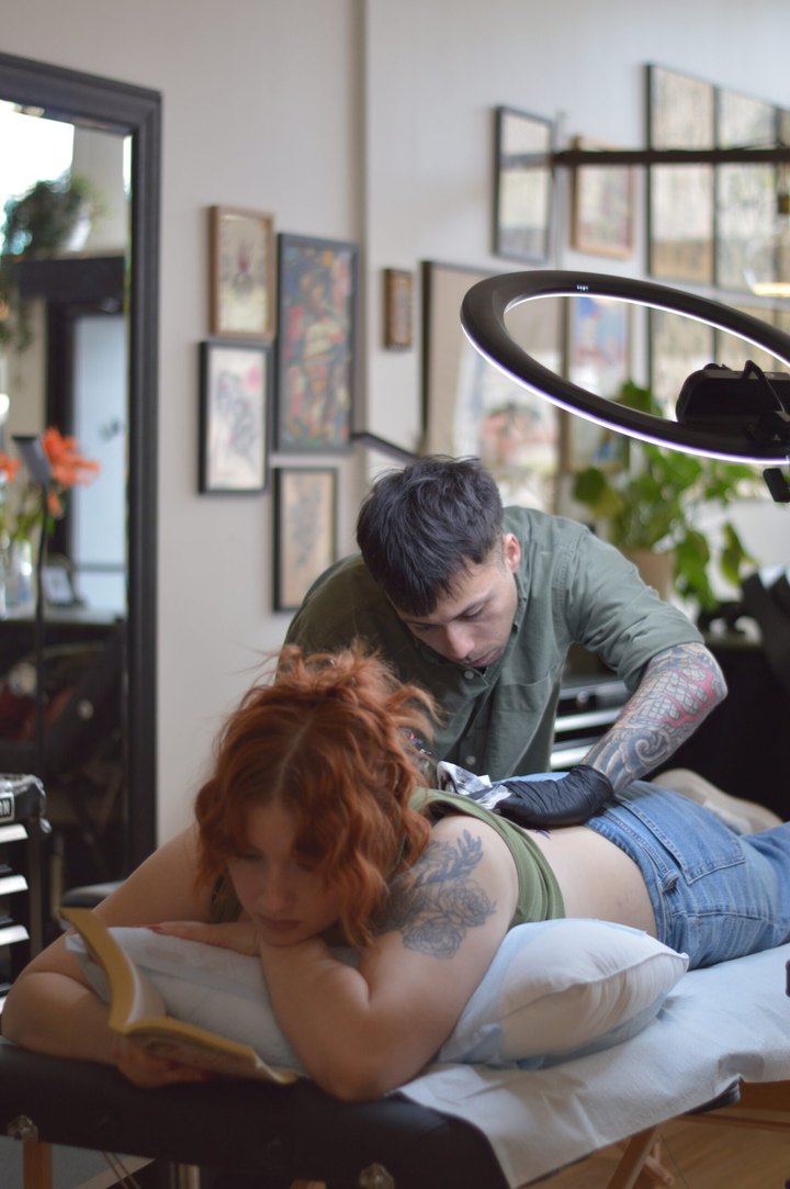 An artist working on the upper back of a client who is lying down reading a book.