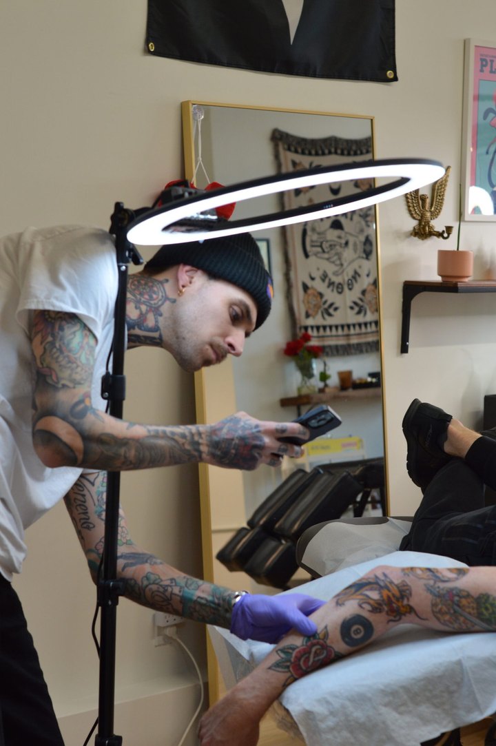 A gloved artist mid-session, working on a client's already-tattooed lower leg.