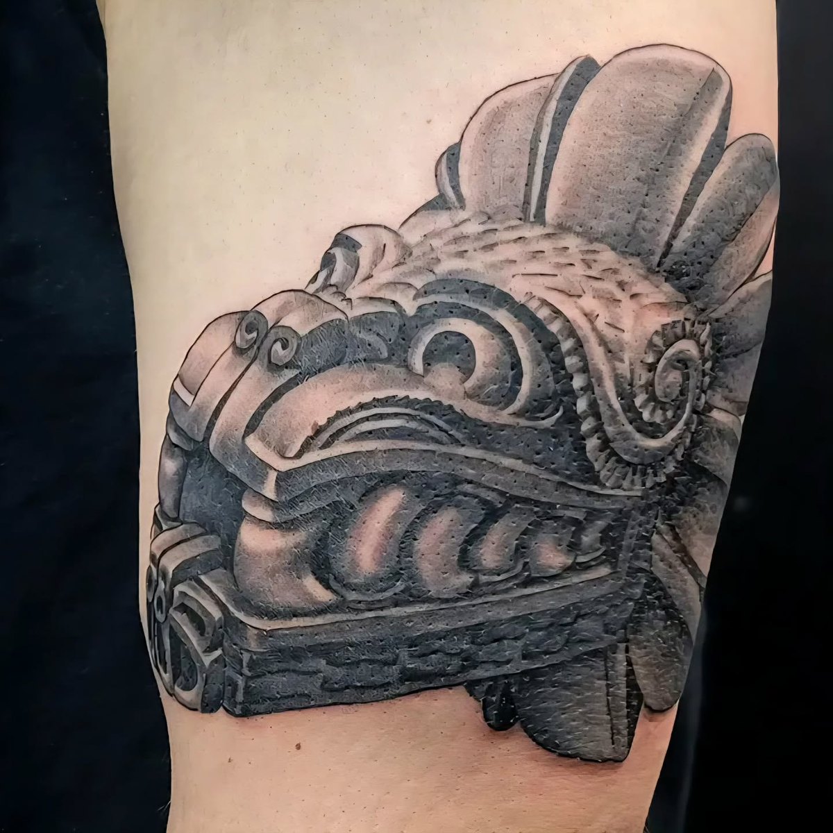 A healed black and grey Aztec feathered-serpent stone carving tattooed on an upper arm.