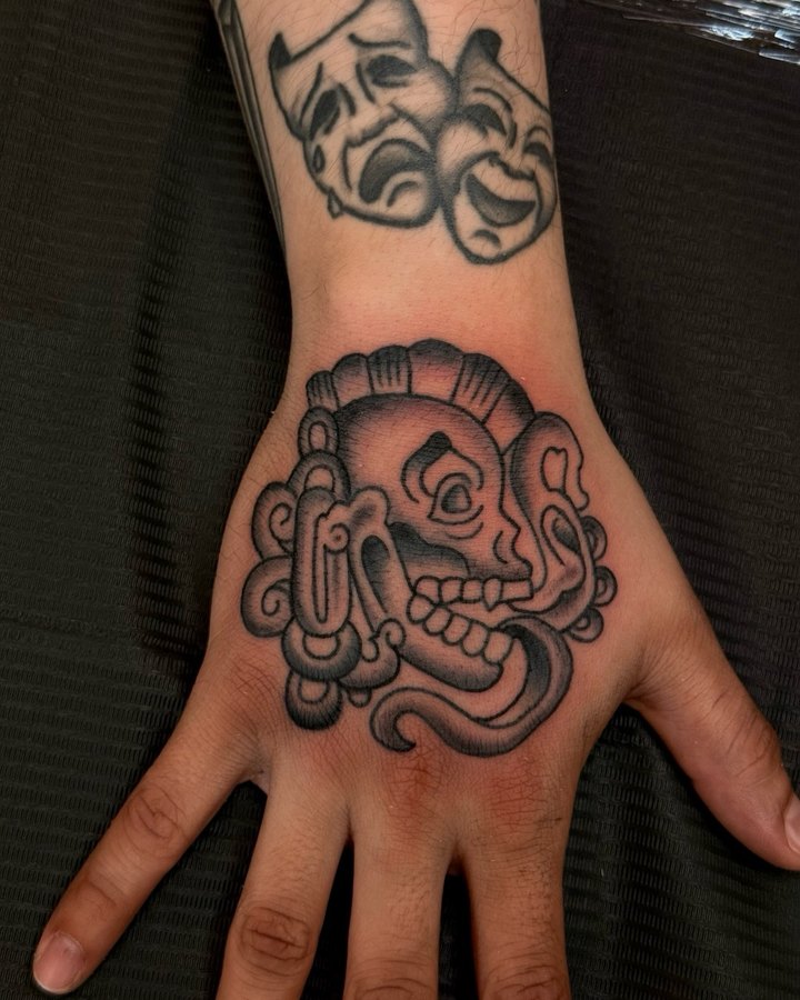 A black and grey Aztec deity head tattooed on a hand.