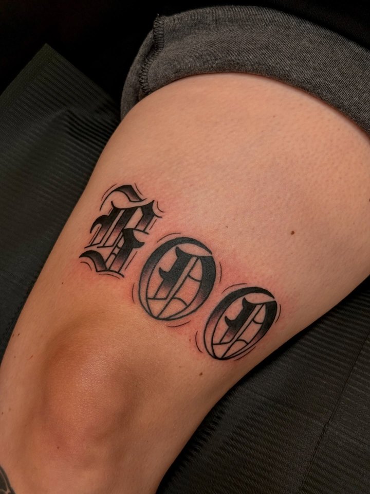 The word BOO in shaded old English blackletter tattooed on a thigh