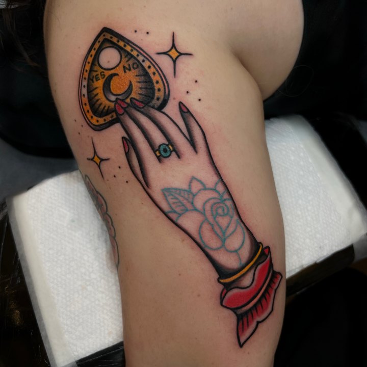 An American traditional hand holding an ouija planchette and a dagger, tattooed on an upper arm.