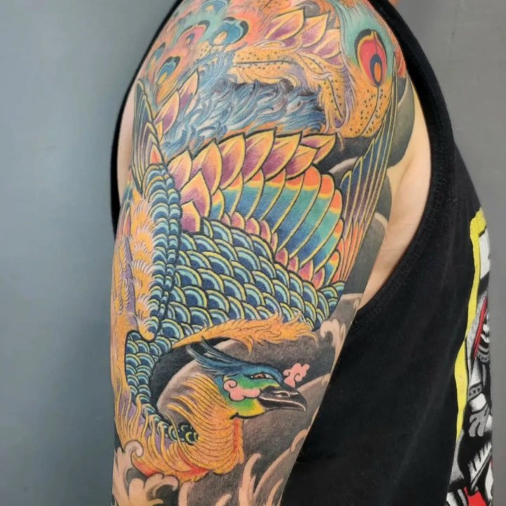 A full colour Japanese phoenix sleeve in orange, blue and teal on an upper arm.
