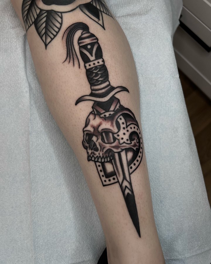 A dagger run through a skull, tattooed on a calf in black and grey